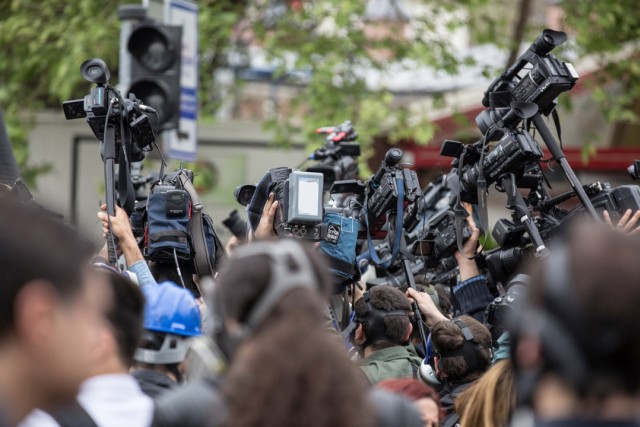 Journalists raise cameras during a crowded outdoor press event.