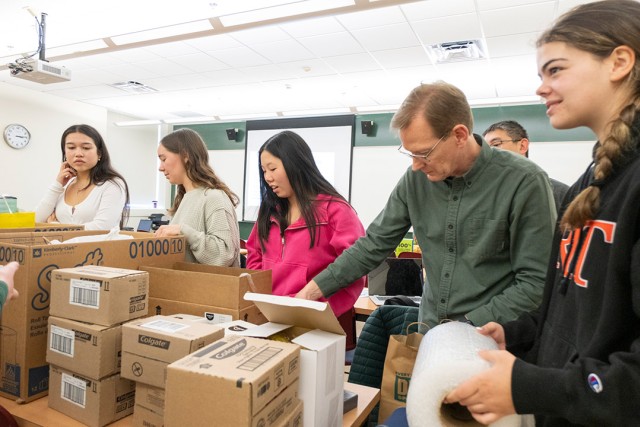  group of students and an instructor sort and organize donation supplies in a classroom, surrounded by cardboard boxes and packing materials.