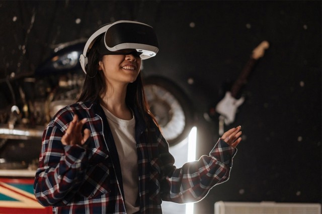 Smiling woman wearing a VR headset and plaid shirt, raising her hands while immersed in virtual reality. A motorcycle wheel and a guitar hang on the dark wall behind her.