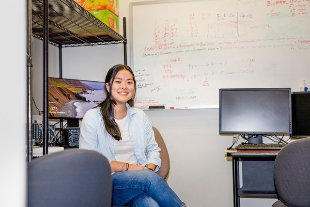 a college age woman in a light blue shirt sits in an office space in front of a whiteboard and shelving unit.