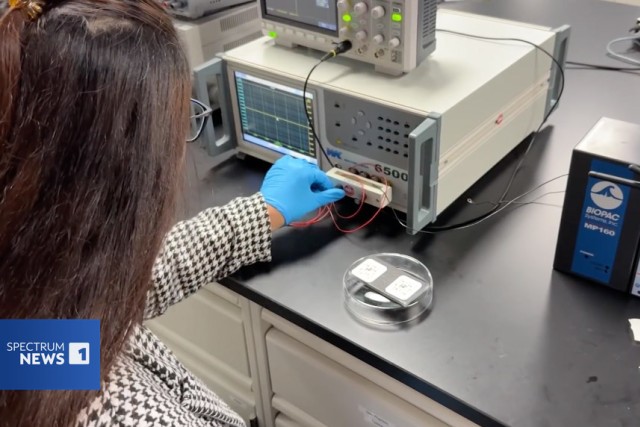 Researcher connects wires to lab equipment while monitoring data on a screen.