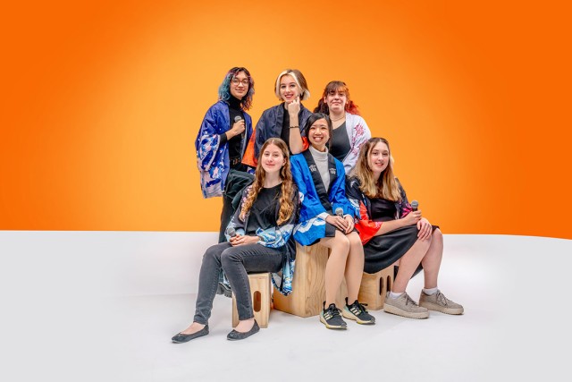 Six young women wearing colorful kimono-style jackets pose confidently in front of an orange backdrop.