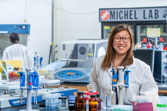 Smiling scientist in lab coat standing in a laboratory with equipment and supplies.
