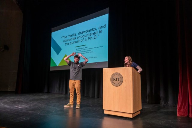 a young female stands behind an R I T podium on stage with a presentation displayed on screen next to her. A male stands next to the podium with his hands in the air gesturing.