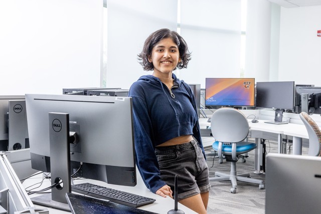 A young woman in a blue sweatshirt and black shorts stands in a computer lab.