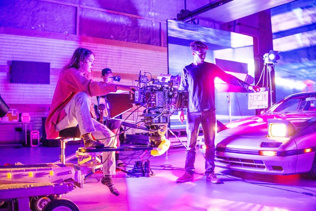 Film set with a person operating a camera and another holding a clapboard near a lit sports car.