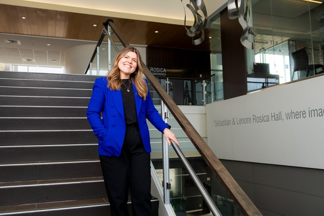 Woman in a blue blazer smiles while standing on stairs inside Sebastian & Lenore Rosica Hall.