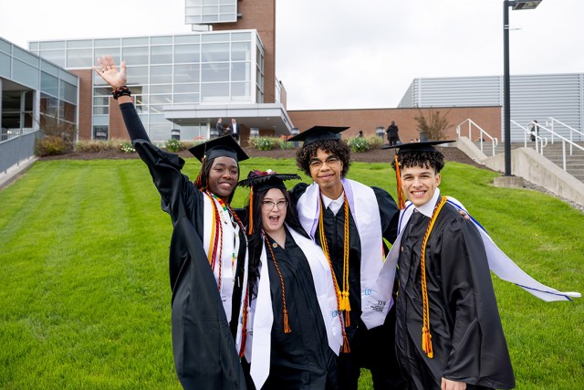 Four graduates in caps and gowns celebrating on a grassy area in front of a modern building.
