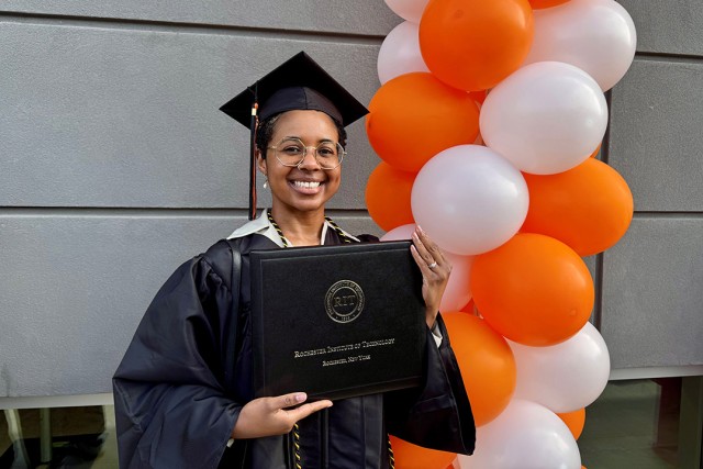 a woman stands in front of a grey cement building and next to orange and white balloons in graduate robes holding a diploma.