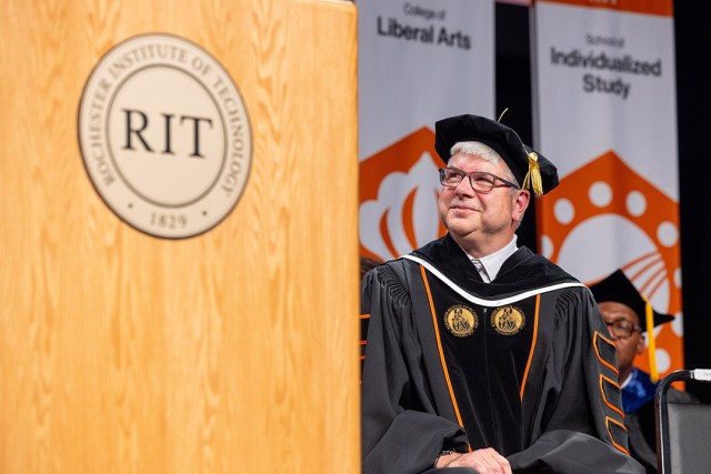 R I T President Bill Sanders stands on a stage and looks happily at the R I T logo