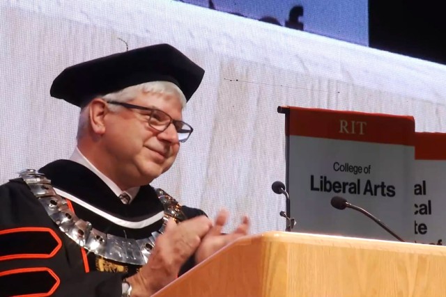 Bills Sanders stands clapping at a podium on a stage at R I T.