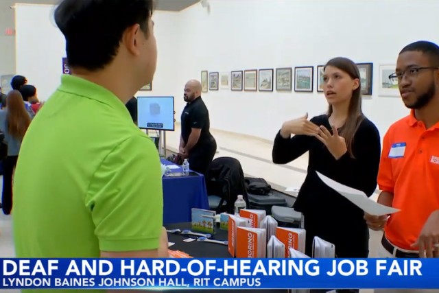 A screenshot of the video news cast. Along the bottom of the image on a blue banner reads deaf and hard-of-hearing job fair, Lyndon Baines Johnson Hall, RIT Campus. On the video there are three people. One man stands in a lime green shirt facing away from the camera. He is talking to a man and a woman standing behind a career fair booth. The man and woman are staring toward the man and the camera engaged in conversation with him.