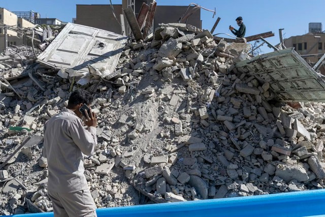 Rubble from a police station damaged in airstrikes on March 3, 2026 in Tehran, Iran. A man in the foreground is seen with his back turned talking on the phone. Another man stands on the top of the pile of rubble taking photos with his phone.