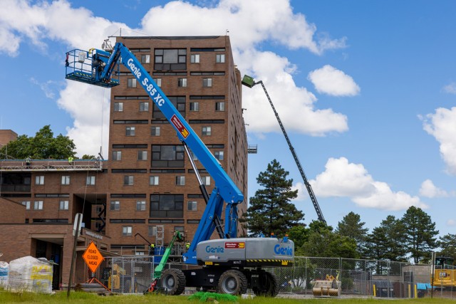 a blue crane with a bucket attachment holds a man high atop a residence hall. More construction equipment is around and in the background.