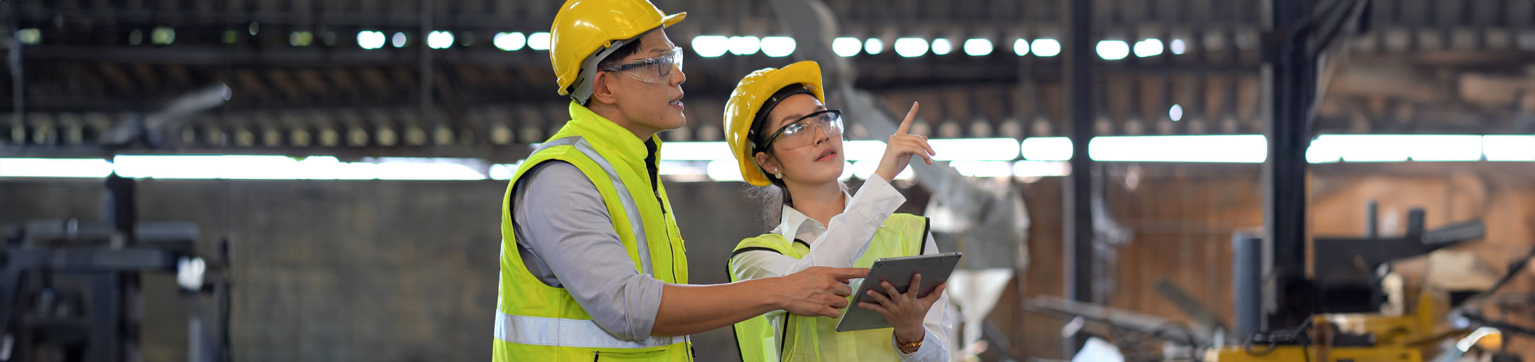 A man and woman in hardhats looking at a tablet and pointing in a manufacturing building.