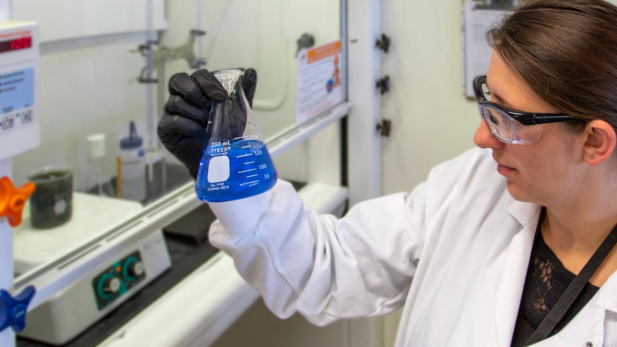 woman in a white lab coat wearing safety glasses and black gloves holding a beaker with blue liquid
