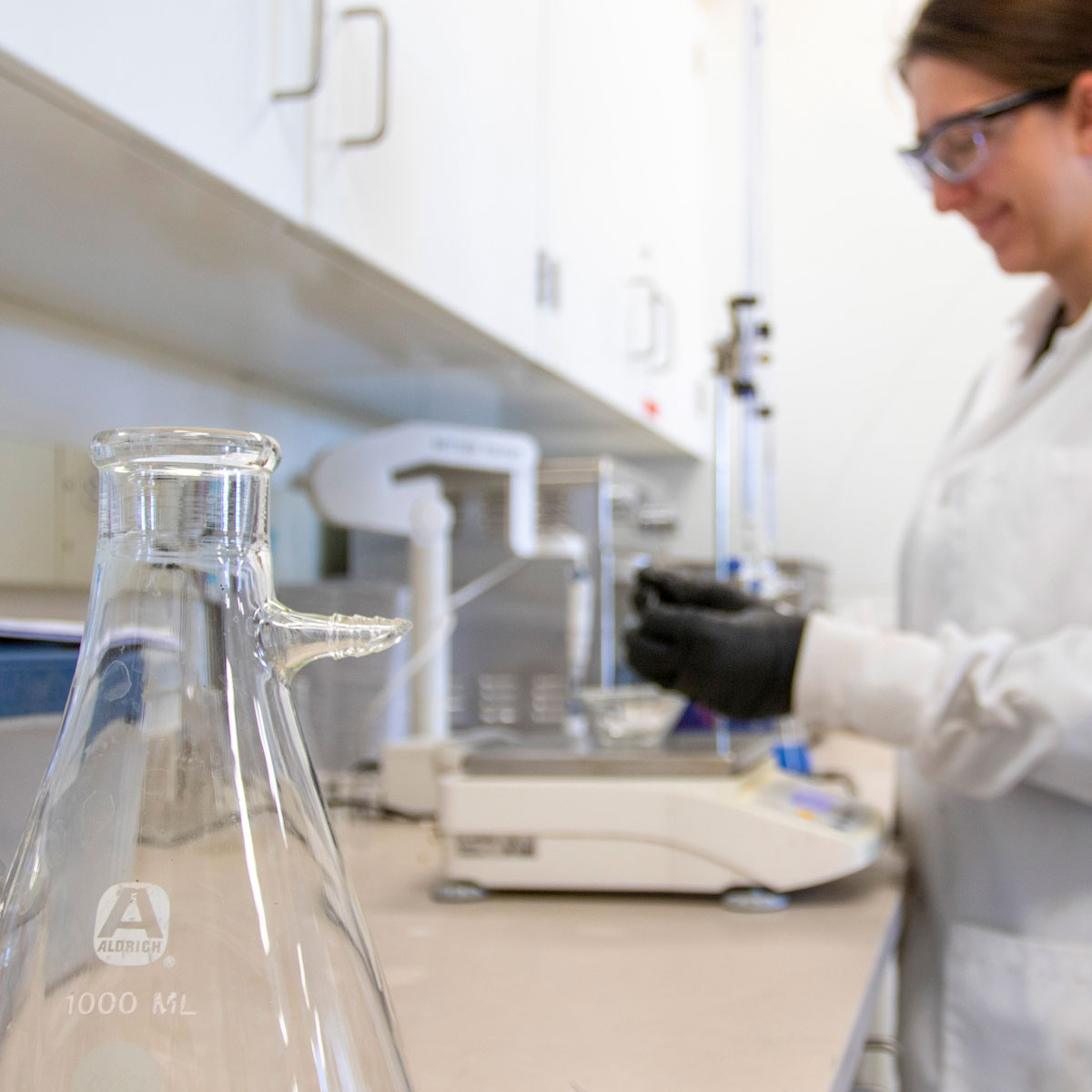 close up of an empty beaker with a blurred image of a woman working in the background smiling and wearing a white lab coat