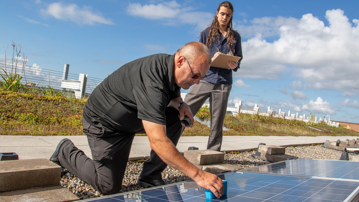 one man kneeling taking measurement on a solar panel while another man is standing behind him with a clipboard