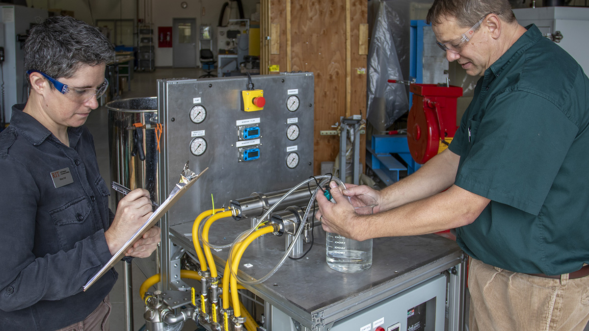 Woman with a clipboard and man holding a tube and taking a measurement inside a large beaker on a large metal device with yellow tubes