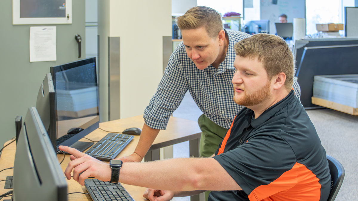 a man sitting at a desk pointing to a computer screen while a woman looks over his shoulder with another worker blurred in the background