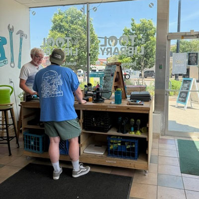 looking from behind the counter at a two men conversing in the front of the Tool Library store