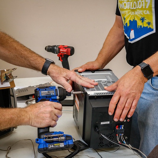 two people working to fix a toaster as one holds it an another uses and electric drill to remove screws