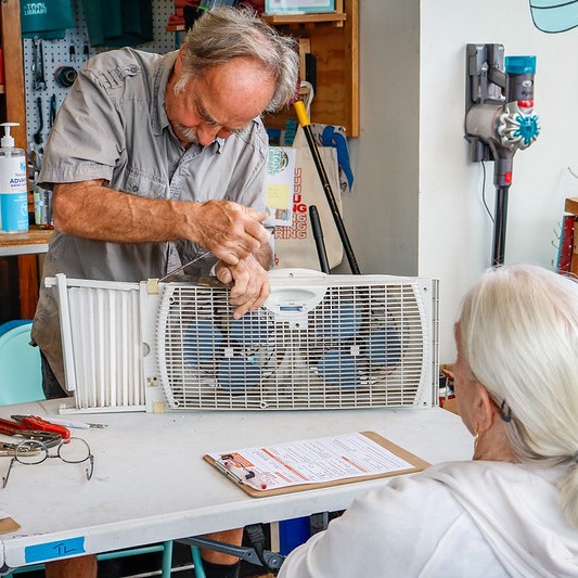 a man fixing a white window fan on a table while a woman watches