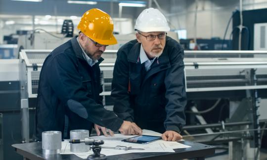 Two people in hardhats looking over plans on paper and a tablet, in a manufacturing building.