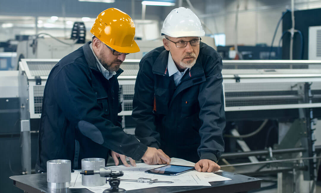 Two people in hardhats looking over plans on paper and a tablet, in a manufacturing building.