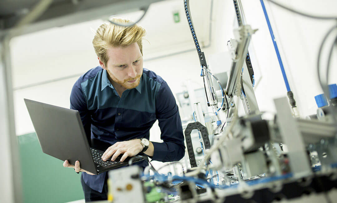 A worker with a laptop inspecting a piece of machinery.