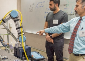A professor pointing at a robotic arm as a student watches.