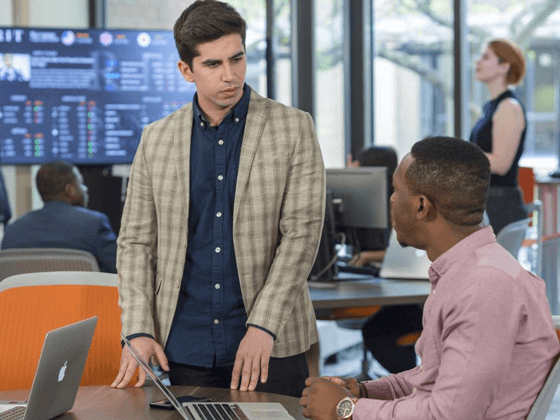 Students sit in front of their laptops in a lab space, with another person standing at the table with them.