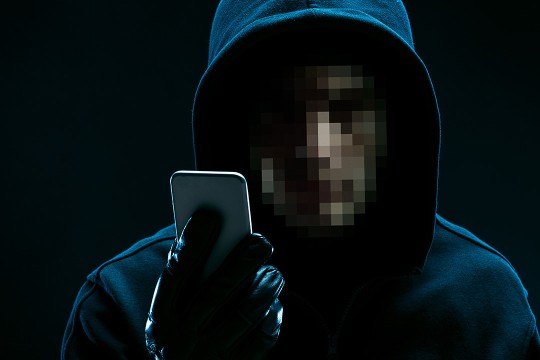 person with a pixelated face wearing a dark hoodie and gloves looking at a cellphone. 