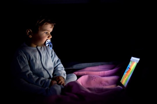 A child with a pacifier stares at a screen in the dark.