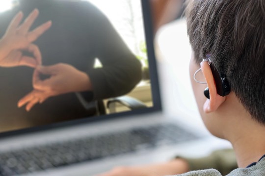 Showing a view of looking over someone's shoulder, the photo features a laptop with a video of someone using ASL. In the foreground, a person with a hearing aid watches the screen. Only the side of their face and hair are visible as the view is looking over their shoulder. 