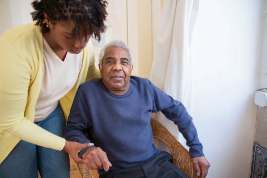 an older man in a rattan chair is being helped up by a younger woman in a yellow sweater