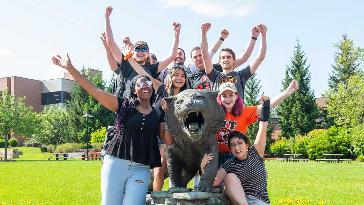 Nine RIT students crowd around a tiger statue and cheer with their arms in the air.