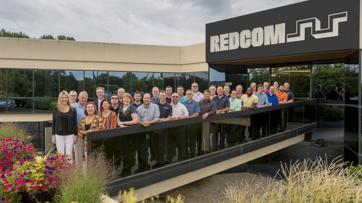 A group of people standing outside of a building with the sign Redcom displayed.