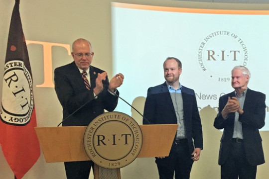 Person clapping standing behind a podium, accompanied by two other people.