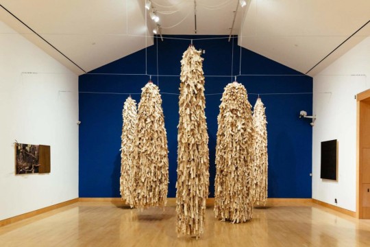 five large sculptures made of layers of paper tags suspended from the ceiling.