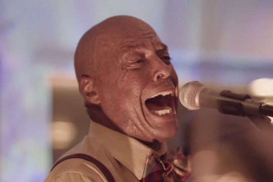 screengrab from a film in which a man is singing into a microphone.