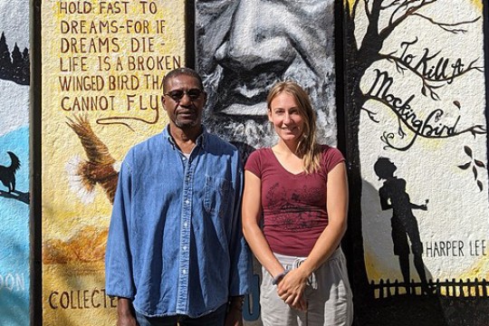 Two artists stand in front of a wall mural inspired by book titles.