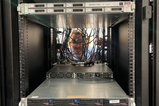 A person wearing glasses behind a server rack peeks through a tangle of cords.