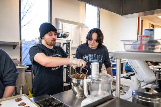 two college students working in a kitchen, scraping batter off of a mixing paddle.