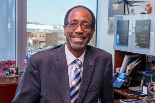Clayton Turner is shown wearing glasses and sitting in his office that looks out over NASA Langley Research Center. He is wearing a gray suit and a blue and white plaid tie.