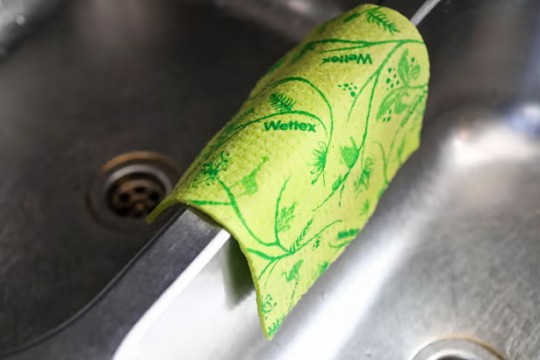 a green swedish dishcloth is shown hanging over the side of a stainless steel sink to dry.