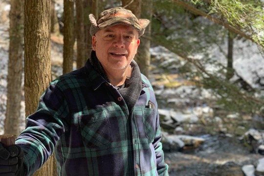 A man in plaid jacket stands outside with trees and a stream in the background