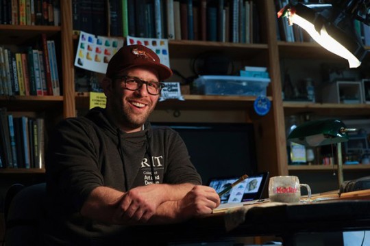 Nick Brandreth sits at a drafting table holding a pencil.