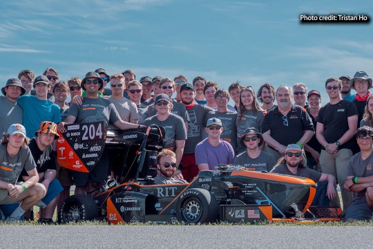 The R I T Formula Racing team stands together iwith their car outside.