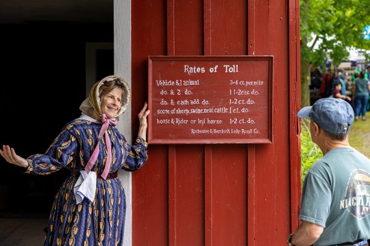 a woman in period dress stands in the doorway of a red building at Genesee Country Village and Museum, showing a sign for Rates of Toll to a male visitor.